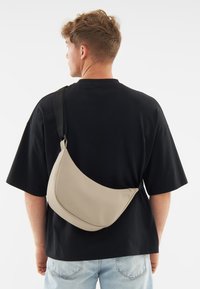 Beige válltáska sima bőrhatású anyagból, ívelt formával és vastag, állítható fekete pánttal.
