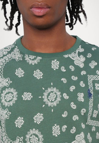 Groen katoenen T-shirt met witte paisley- en bloemenpatronen. Ronde hals, korte mouwen, met een klein blauw logo op de borst.