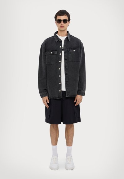 Zwarte denimjas met twee borstzakken, witte t-shirt, zwarte shorts en witte sneakers. Model draagt zwarte zonnebril en enkelsokken.