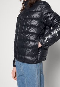 Svart, vadderad pufferjacka med en glansig finish, hel dragkedja, hög krage, sidofickor och logotypdetalj på ärmen.
