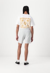 Hvit t-skjorte med oransje blomsterkvadratgrafikk på baksiden, kombinert med grå shorts. Modellen har på seg svarte joggesko med hvite såler.
