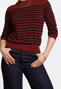 Femme portant un pull à rayures rouille et noir à manches longues, un jean bleu foncé, un collier en chaîne dorée, un bracelet et des bagues, les mains dans les poches.