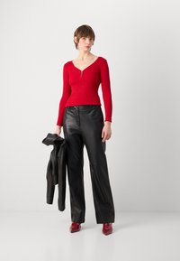 Haut rouge à manches longues à côtes avec un détail de fermeture éclair, associé à un pantalon large en cuir noir et des escarpins rouges à bout pointu. Veste en cuir noir portée sur le bras.