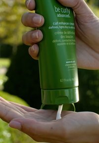 Groene plastic fles met krulversterker-crème die een witte substantie in een handpalm doseert; bevat tekst die de voordelen van het product en het volume beschrijft.