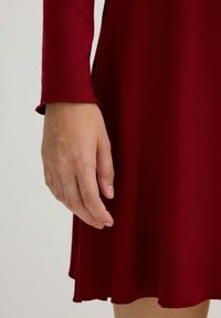 Vestido burdeos con mangas largas y dobladillo acampanado. La tela es suave con un ligero brillo. Los extremos de las mangas tienen un sutil detalle de dobladillo enrollado.