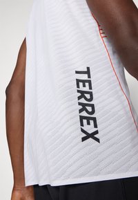 Biała bezrękawnikowa bluzka z teksturowanym wzorem, podkreślona pomarańczowym szwem oraz dużym czarnym logo "TERREX" po boku.