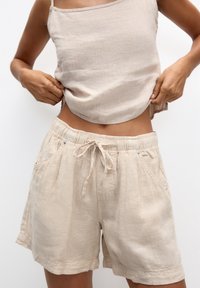 Top senza maniche in lino beige e pantaloni corti abbinati. I pantaloni corti presentano una vita elasticizzata, cordoncino e due tasche laterali. Texture liscia.
