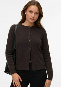 Brūns adīts kardigans ar garām piedurknēm, apaļu kakla izgriezumu un pogu aizdari. Kombinēts ar melnu teksturētu somiņu. Gluda un silta tekstūra.