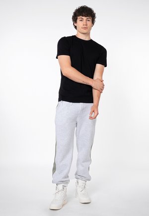 Jonge man staand, draagt een zwart T-shirt, lichtgrijze joggingbroek met zijstrepen, en witte sneakers, houdt zijn linkerarm vast met zijn rechterhand.