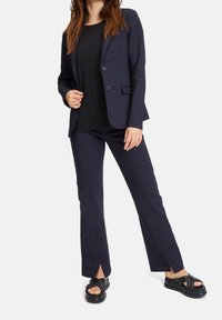 Marinblå skräddarsydd kavaj med två knappar, kombinerad med en veckad svart topp och figurnära marinblå byxor med sidslitsar. Svarta platåsandaler.