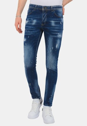 Slim-fit blauwe versleten spijkerbroek met vervaagde plekken en verfspatten, gedragen met witte sneakers en een wit overhemd.