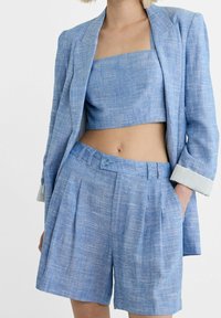 Blazer di lino azzurro chiaro su un crop top coordinato e pantaloni corti plissettati. Il blazer presenta maniche a risvolto e una vita con bottone.