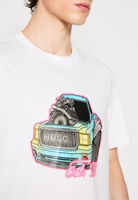 Bílý bavlněný tričko s ilustrací barevného kamionu s detaily motoru, štítkem "HUGO" a nápisem "Est. 93" v růžovém textu.