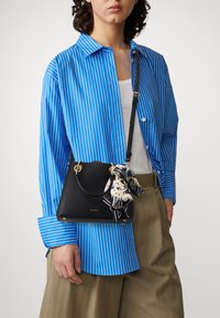 Black leather handbag with a textured finish, featuring gold hardware and a patterned scarf attached. Model wears a blue striped shirt and beige pants.