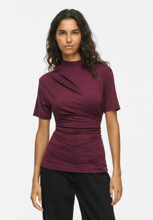Femme aux longs cheveux bouclés porte un haut bordeaux ajusté à manches courtes et un pantalon noir, debout devant un fond blanc uni.