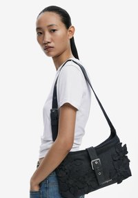 Black fabric shoulder bag with floral appliqué and silver buckle detail, featuring a wide strap and a structured shape.