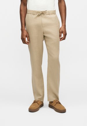 Άνδρας που φοράει μπεζ λινά παντελόνια με κορδόνια και καφέ σουέτ παπούτσια, στέκεται μπροστά σε ένα απλό λευκό φόντο.