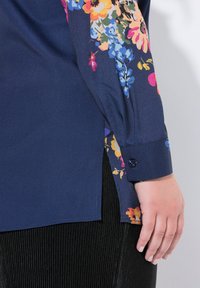 Marineblaue Bluse mit bunten Blumenmustern an der Seite und dem Ärmel, langen Ärmeln, Manschette mit Knopfdetail, glatter Textur.