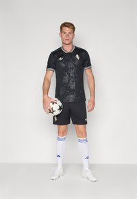 Maillot de football noir avec un imprimé graphique, bordures vertes, short noir et chaussettes blanches à rayures bleues. Tenant un ballon de football noir et blanc.