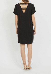 Svart kortärmad klänning med V-formad rygg och geometriska urtag, knälång, slät textur, ihop med beige strap-on klackar.
