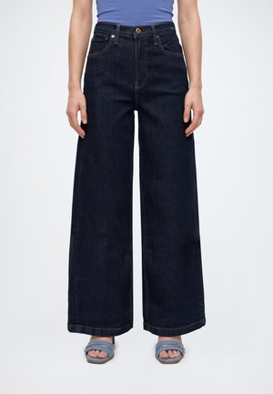 Un mannequin se tient devant un fond blanc uni, portant un jean indigo foncé taille haute à jambes larges, avec des poches avant, et des sandales bleues.