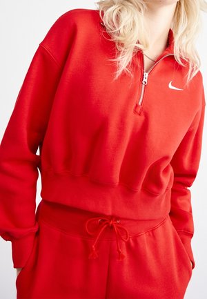 Kobieta w czerwonej bluzie Nike z kołnierzem na zamek i dopasowanych czerwonych spodniach ze sznurkiem, stojąca z rękami w kieszeniach.