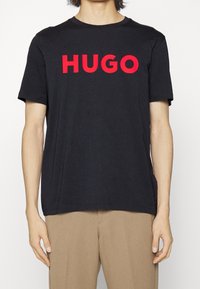 Černé bavlněné tričko s nápisem "HUGO" vytištěným tučnými červenými písmeny. Krátké rukávy, kulatý výstřih a volný střih. Jednoduchý design bez vzorů.