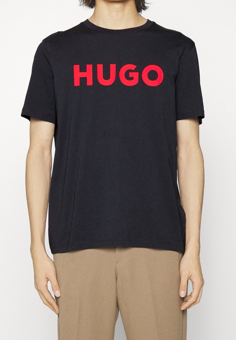 Černé bavlněné tričko s nápisem "HUGO" vytištěným tučnými červenými písmeny. Krátké rukávy, kulatý výstřih a volný střih. Jednoduchý design bez vzorů.