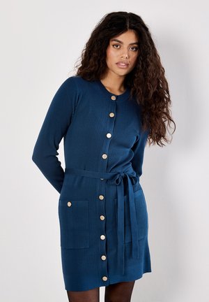 Mujer con cabello rizado y largo, vestida con un vestido de punto acanalado azul, con botones, bolsillos frontales y un cinturón anudado en la cintura, de pie frente a una pared blanca.