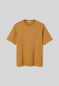 T-shirt jaune moutarde à manches courtes et col rond avec un petit logo noir et blanc sur la poitrine gauche sur fond gris.