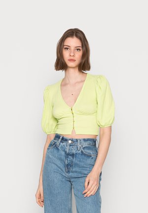 Blusa cropped verde-claro com mangas bufantes, decote em V e seis botões decorativos. Usada com jeans de ganga azul desbotada e de cintura alta.