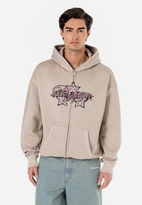 Beige zip-up hoodie with large graphic print in pink and black, featuring a front pouch pocket and relaxed fit. Soft fabric, long sleeves.