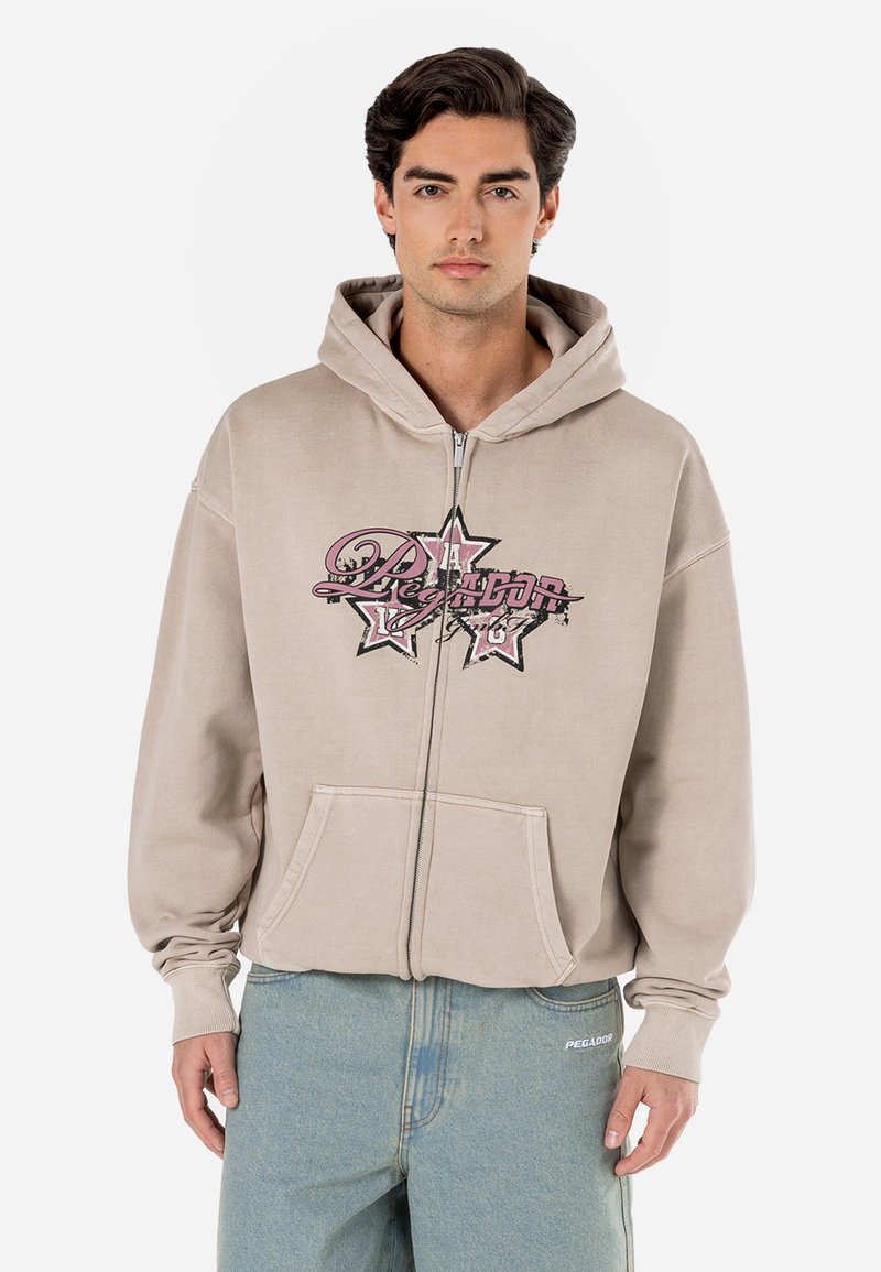 Beige zip-up hoodie with large graphic print in pink and black, featuring a front pouch pocket and relaxed fit. Soft fabric, long sleeves.