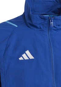 Blaue Jacke mit einem Frontreißverschluss, hohem Kragen, hellblauem Streifen auf der Schulter und weißem Markenlogo mit drei Streifen auf der Brust.