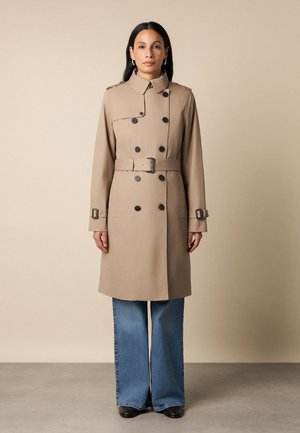 Kobieta stoi zwrócona przodem, ubrana w beżowy płaszcz typu trencz z paskiem i czarnymi guzikami, niebieskie szerokie spodnie dżinsowe oraz czarne buty na jednolitym tle.