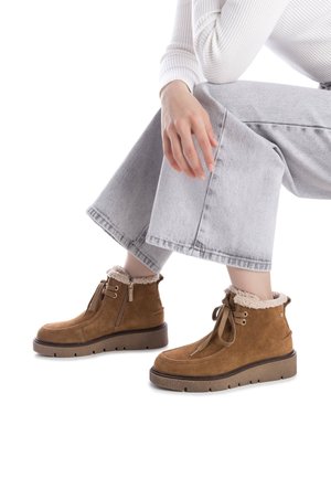 Botas de ante marrón hasta el tobillo con un forro de felpa, suela plana y cremallera lateral. Combinadas con jeans de pierna ancha en gris, mostrando materiales texturizados.