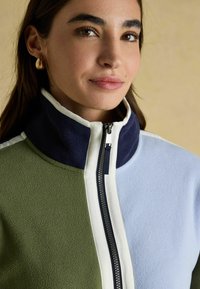 Fleece bunda s farebnými blokmi v olivovo zelenej, námorníckej a svetlo modrej farbe, s vysokým golierom a plným predným zipsom s kontrastným lemovaním.