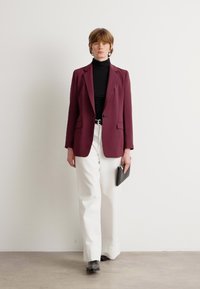 Burgundský blazer přes černý rolák, kombinovaný s bílými kalhotami. Modelka drží černou kabelku a má na sobě černé boty. Minimalistický design.