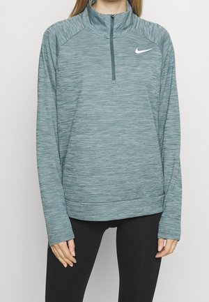 Frau trägt einen langärmligen, grau-grünen Nike-Pullover mit Reißverschluss und schwarze Leggings, steht vor einem einfarbigen hellen Hintergrund.