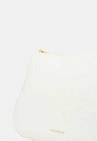 Borsa a mano in pelle bianca con texture a ciottoli, forma arrotondata, dettagli in oro e chiusura con zip. Nome del marchio "COCCINELLE" impresso in oro.