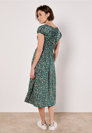 Kobieta w zielonej sukience midi w kwiaty z krótkimi rękawami i marszczonym pasem, w białych trampkach, stojąca bokiem.