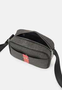 Kompaktní šedá taška přes rameno s černým textovým vzorem, červeným vertikálním pruhem, kovovým logem Valentino a zipem s černým nastavitelným popruhem.