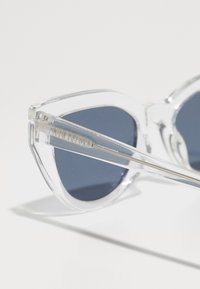 Ochelari de soare tip cat-eye, cu lentile întunecate, având o ramă transparentă și un logo argintiu pe tâmplă. Textură netedă, design modern.