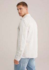 Joven vestido con una camisa de botón color blanco roto y jeans azul claro, mirando sobre su hombro contra un fondo beige liso.