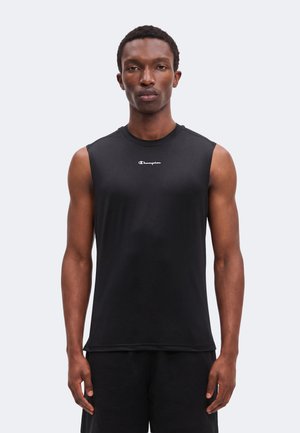 Junger Mann, der ein ärmelloses schwarzes Champion-Shirt und schwarze Shorts trägt, steht vor einem schlichten hellen Hintergrund, frontal zur Kamera gewandt.