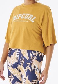 Żółta krótka bluzka z krótkimi rękawami z białym logo "Ripcurl", połączona z granatowymi legginsami z dużymi kremowymi wzorami liści.