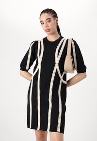 Zwarte gebreide jurk met crèmekleurige haakdetails, korte pofmouwen en een slim fit. Gecombineerd met een kleine beige handtas.