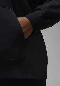 Felpa nera con finitura testurizzata, caratterizzata da una tasca laterale e polsini a costine. Il tessuto appare morbido e flessibile.