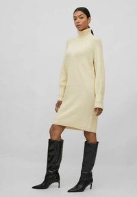 Donna che indossa un vestito in maglia color crema con collo alto e stivali neri con tacco a punta al ginocchio, in piedi con una gamba leggermente avanti.