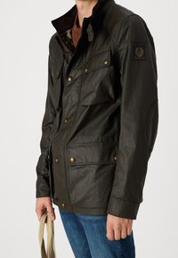 Tmavě zelená voskovaná bavlněná bunda s vysokým límcem, kapsami na knoflíky a bronzovými kovovými detaily. Vhodná na volný čas.